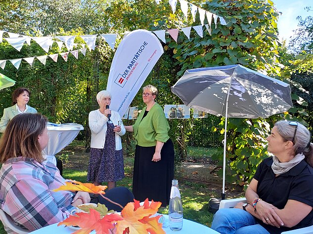 Frau spricht bei einer Gartenveranstaltung vor sitzenden Gästen.