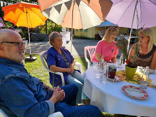 Vier Personen sitzen unter bunten Sonnenschirmen an einem gedeckten Gartentisch.