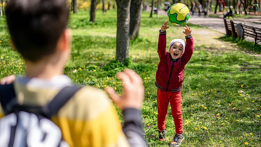 Kind fängt einen grünen Ball im Park, im Vordergrund unscharf ein weiteres Kind