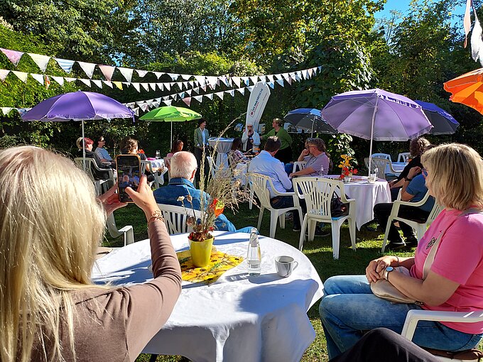 Gartenfest mit Gästen an gedeckten Tischen, Sonnenschirmen und Wimpelketten.