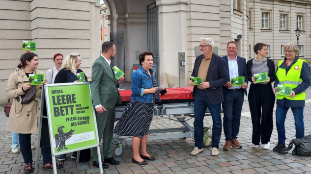 Menschen vor Gebäude mit leerem Klinikbett und grünen Protestplakaten zur Pflege