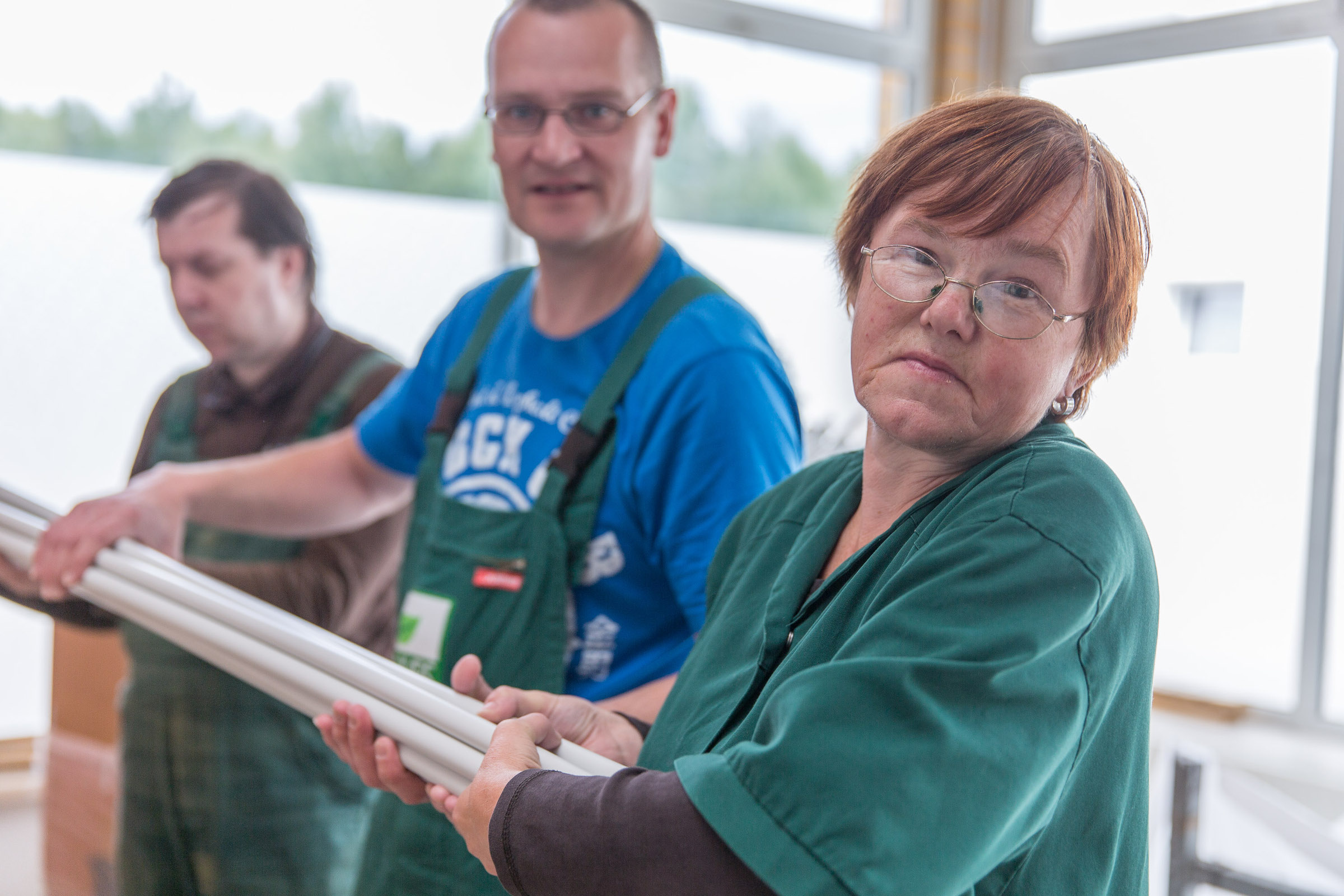  Eine Frau und zwei Männer heben mehrere Stangen gemeinsam auf. Sie tragen Arbeitskleidung und gucken in die Kamera. 
